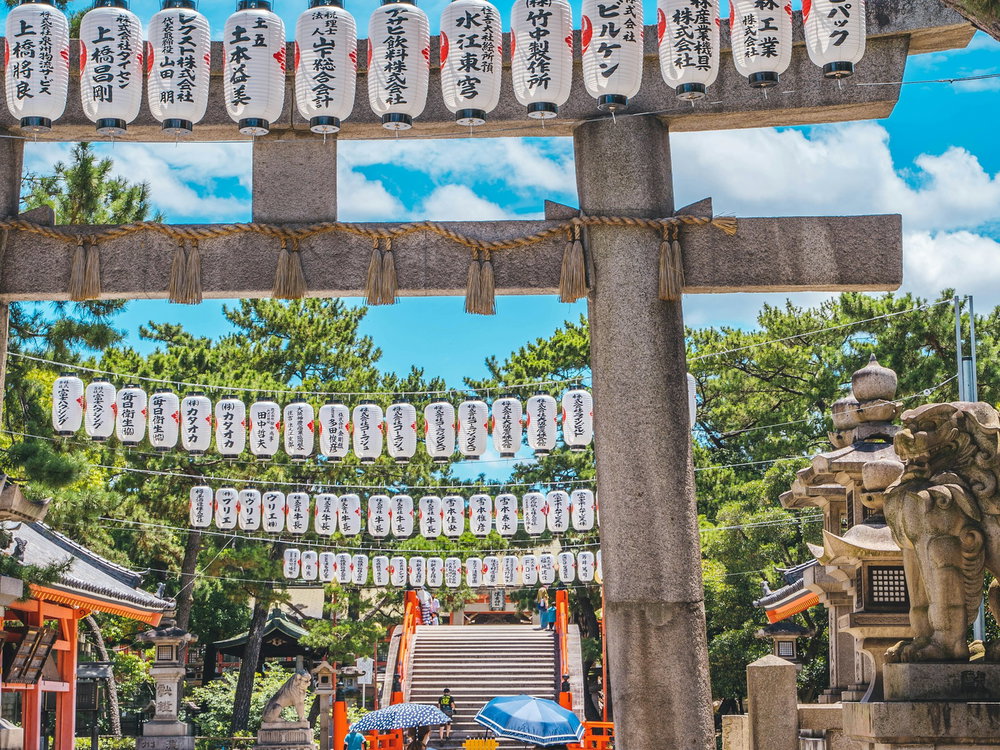 Stone Torii gates with lanterns , stone statuesand bright sky with green trees, two women carrying umbrellas 