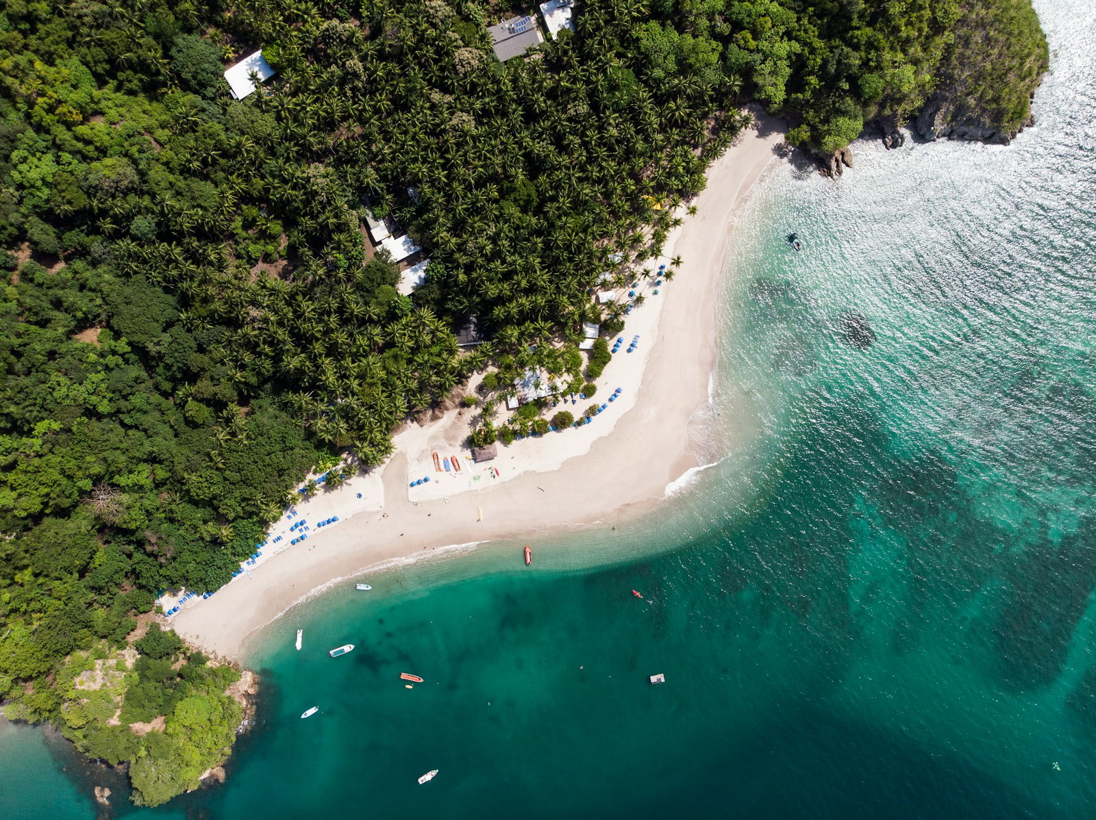 Deep turquoise ocean with some boats that curves around white sand beach and green jungle with a few houses, aerial shot