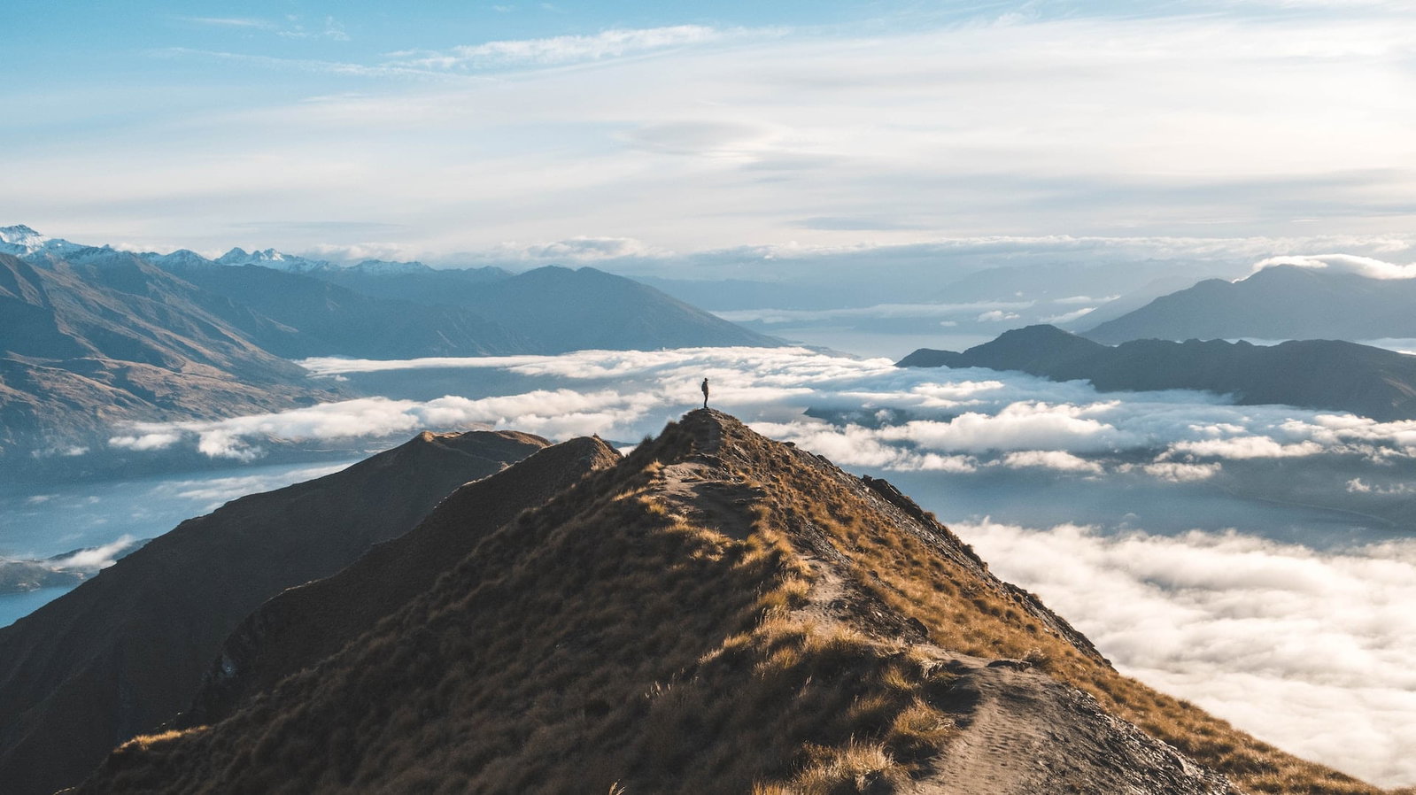 A lone traveller stands on top of a mountain with a blanket of clouds below