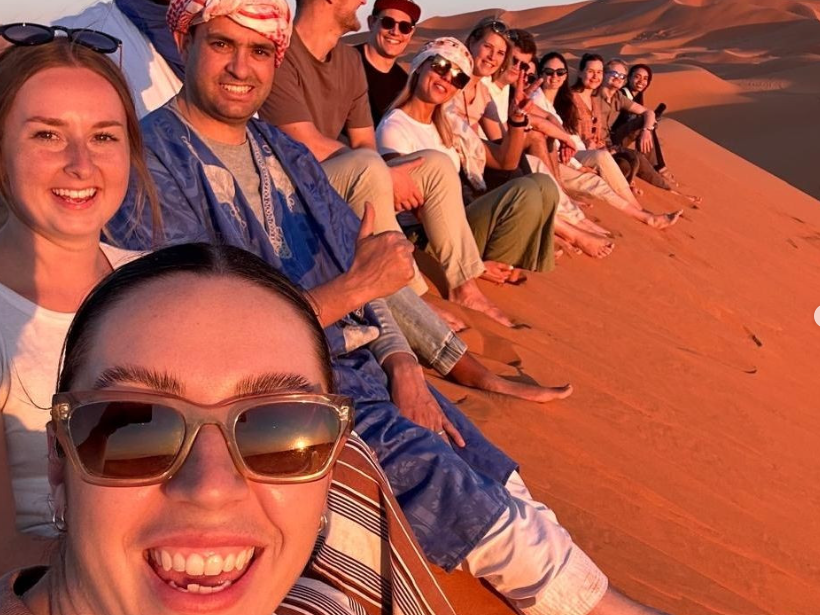 Group of travellers sat at the top of a sand dune in the Sahara Desert at sunset in Morocco